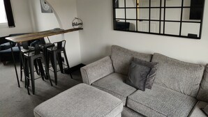 A living room with a grey sofa, a wooden dining table with chairs, a black pendant light, and a large mirror on the wall. - 1 Bedroom Riverside Apartment St Neots Riverside Park Area (St Neots)