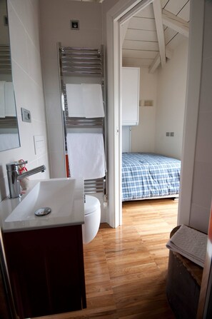 Une salle de bain avec un lavabo, un miroir et un porte-serviettes. La pièce adjacente contient un lit recouvert d’une couverture à carreaux bleus.