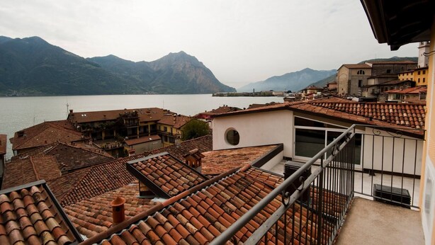 Um terraço com telhas de barro, uma janela redonda e vista para um lago e montanhas.