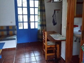 A room with a blue door, a wooden table with a chair, a tiled floor, and a small shelf with items.
