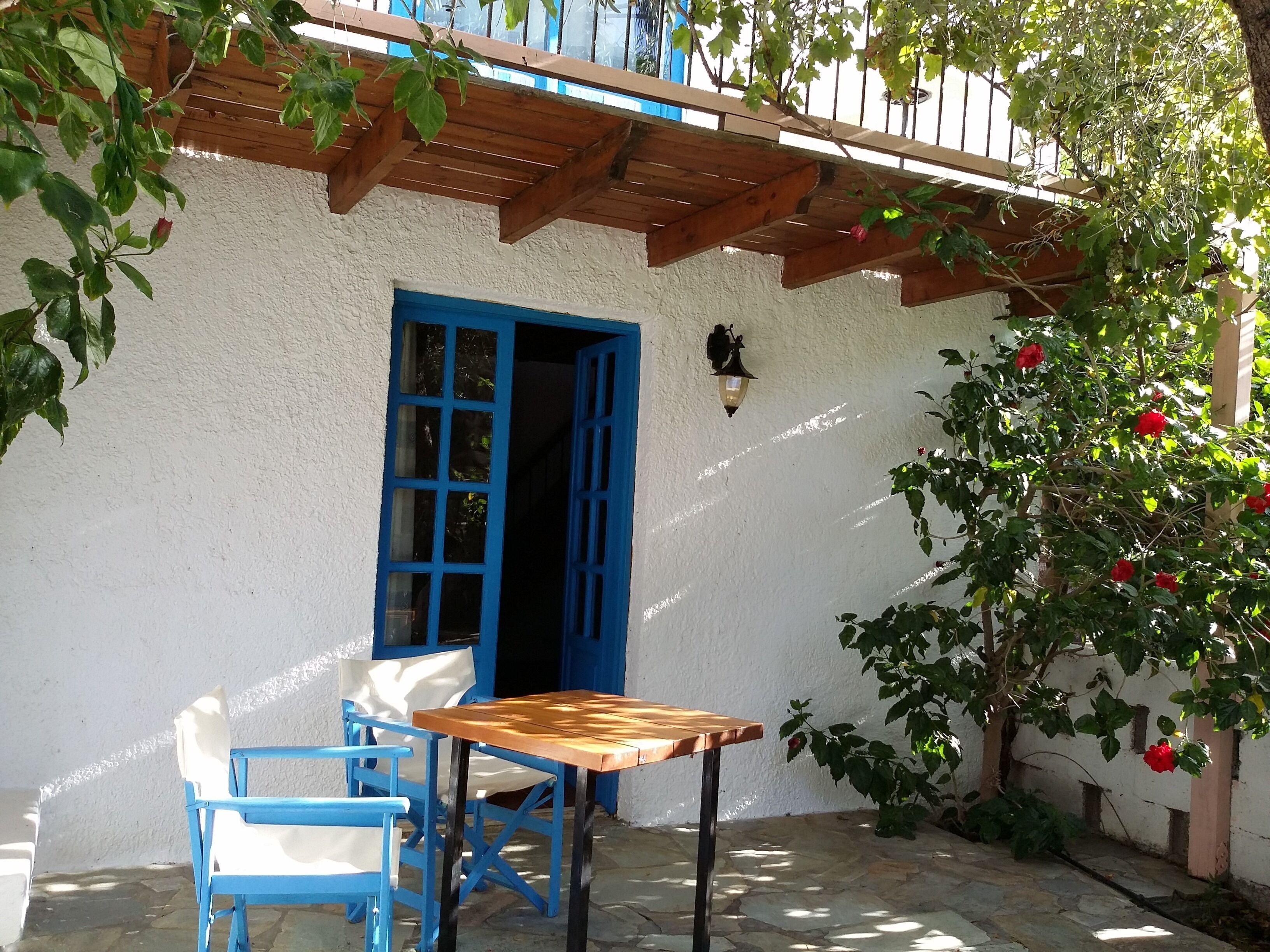 Une terrasse avec une table en bois et des chaises bleues, une porte bleue et un luminaire fixé au mur.