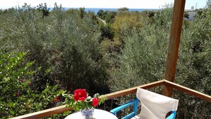 A balcony with a table and blue chairs, overlooking a garden with various plants and a view of the sea.