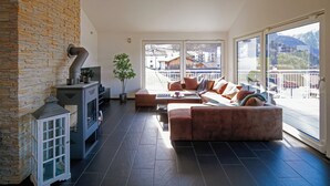 A modern living room with a stone wall, a fireplace, a large window, and a view of buildings and mountains. - Modern and Cosy 5 Rooms Penthouse With Stunning View (Triesenberg)