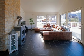 Un salon moderne avec un mur en pierre, une cheminée, un canapé et une vue sur les bâtiments et les montagnes, visible à travers de grandes fenêtres.
