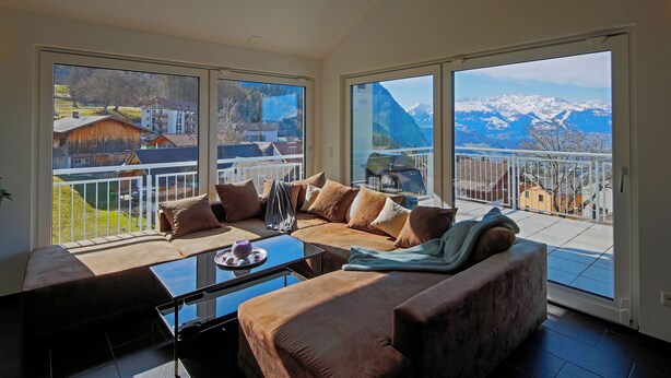 A modern living room with a large sofa, a coffee table, and a view of a mountain landscape through large windows. - Modern and Cosy 5 Rooms Penthouse With Stunning View (Triesenberg)
