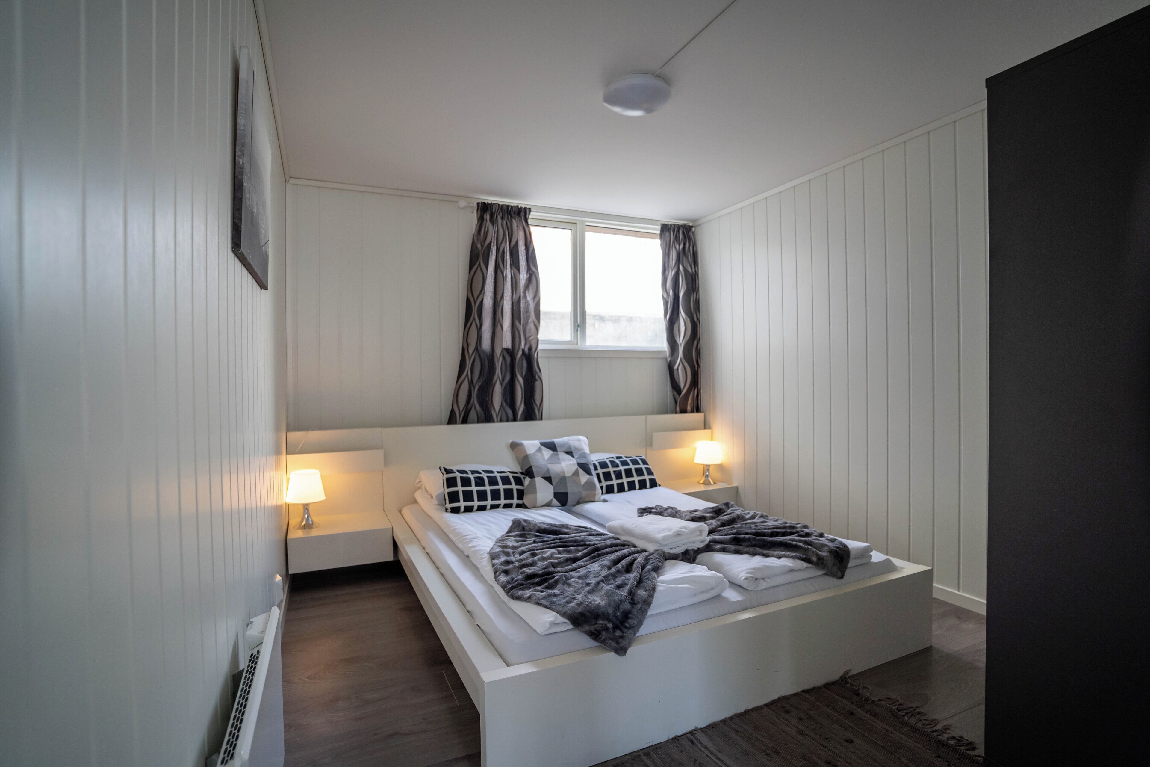 A modern bedroom with a white bed, two bedside lamps, a window with curtains, and a radiator.