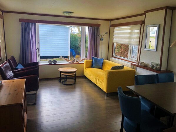 A room with a yellow sofa, a wooden dining table, a small round table, and a window with curtains.