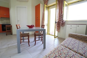 A compact room with a dining table, chairs, a sofa, a kitchen area, and a sliding glass door to the outside.