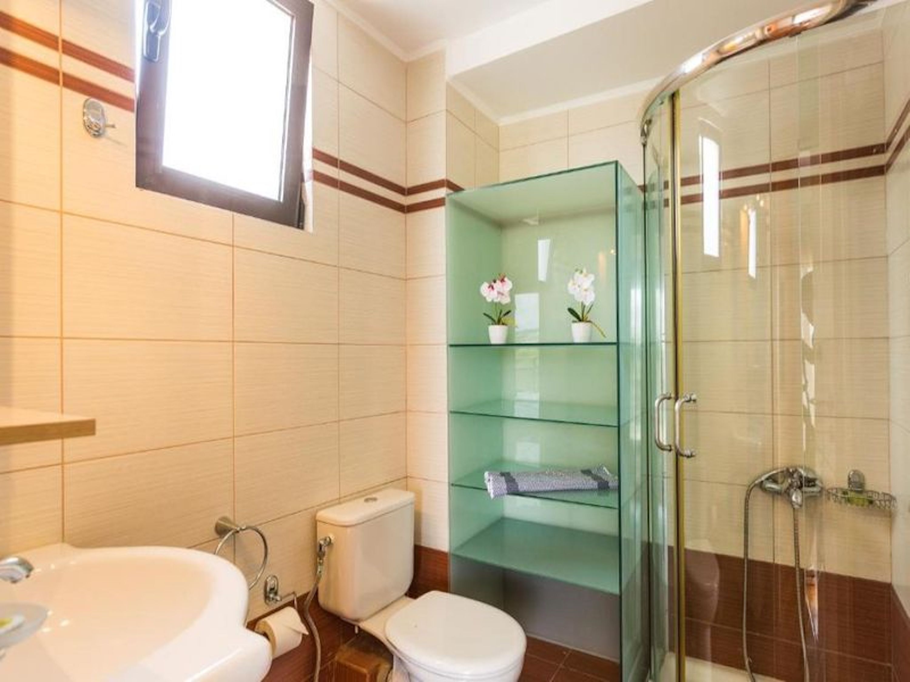 A bathroom with a glass shower enclosure, a toilet, a bidet, and a glass shelf with flowers.