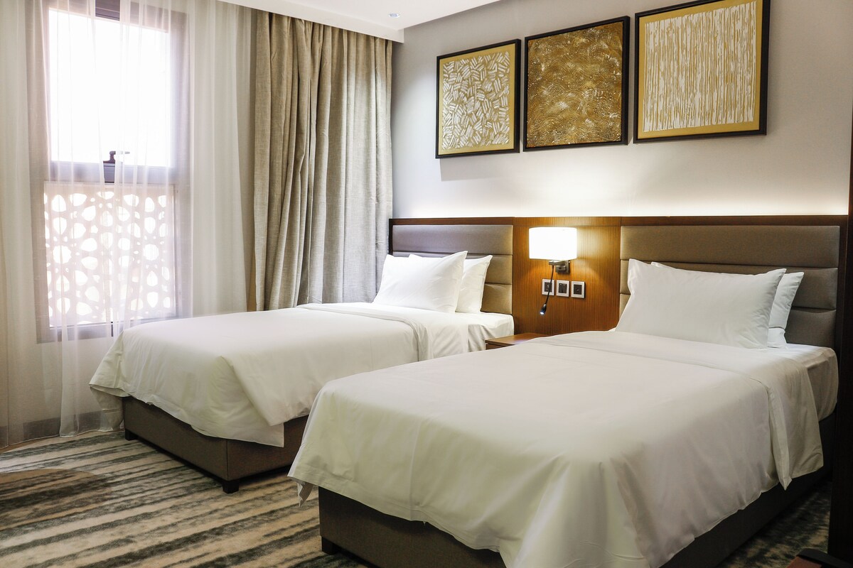 A hotel room with two beds, a large window with curtains, and three framed artworks on the wall.