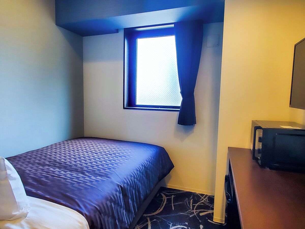 A hotel room with a bed, a small window, a television, and a desk.