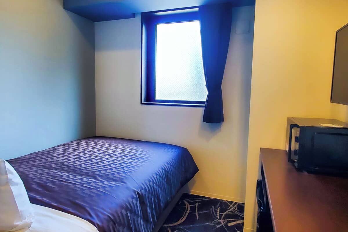Ein Hotelzimmer mit Bett, kleinem Fenster, Fernseher und Schreibtisch.