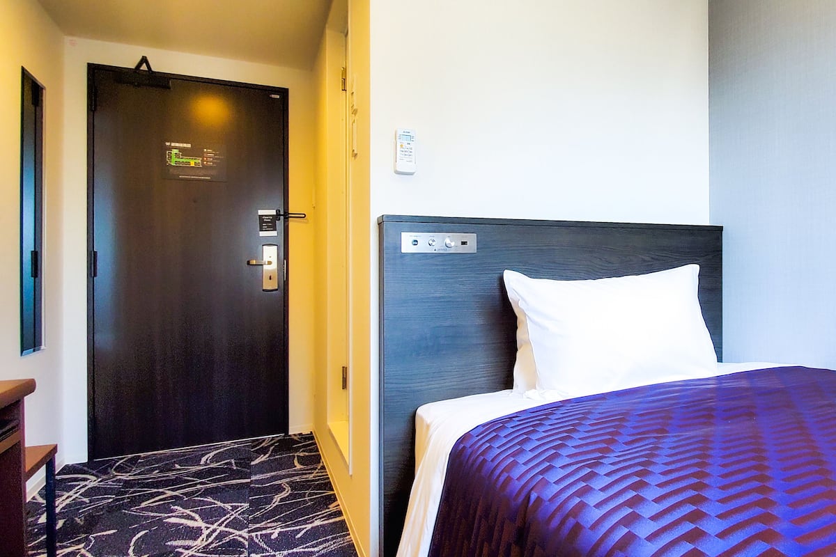 Ein Hotelzimmer mit einer dunklen Holztür, einer Bedienfeld an der Wand, einem Bett mit einem weißen Kissen und einem gemusterten Teppich.
