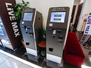 Deux bornes en libre-service, dotées d’un écran numérique et d’un lecteur de cartes, portant l’inscription « LiVEMAX HOTELS & RESORTS », se trouvent dans le hall d’un hôtel.
