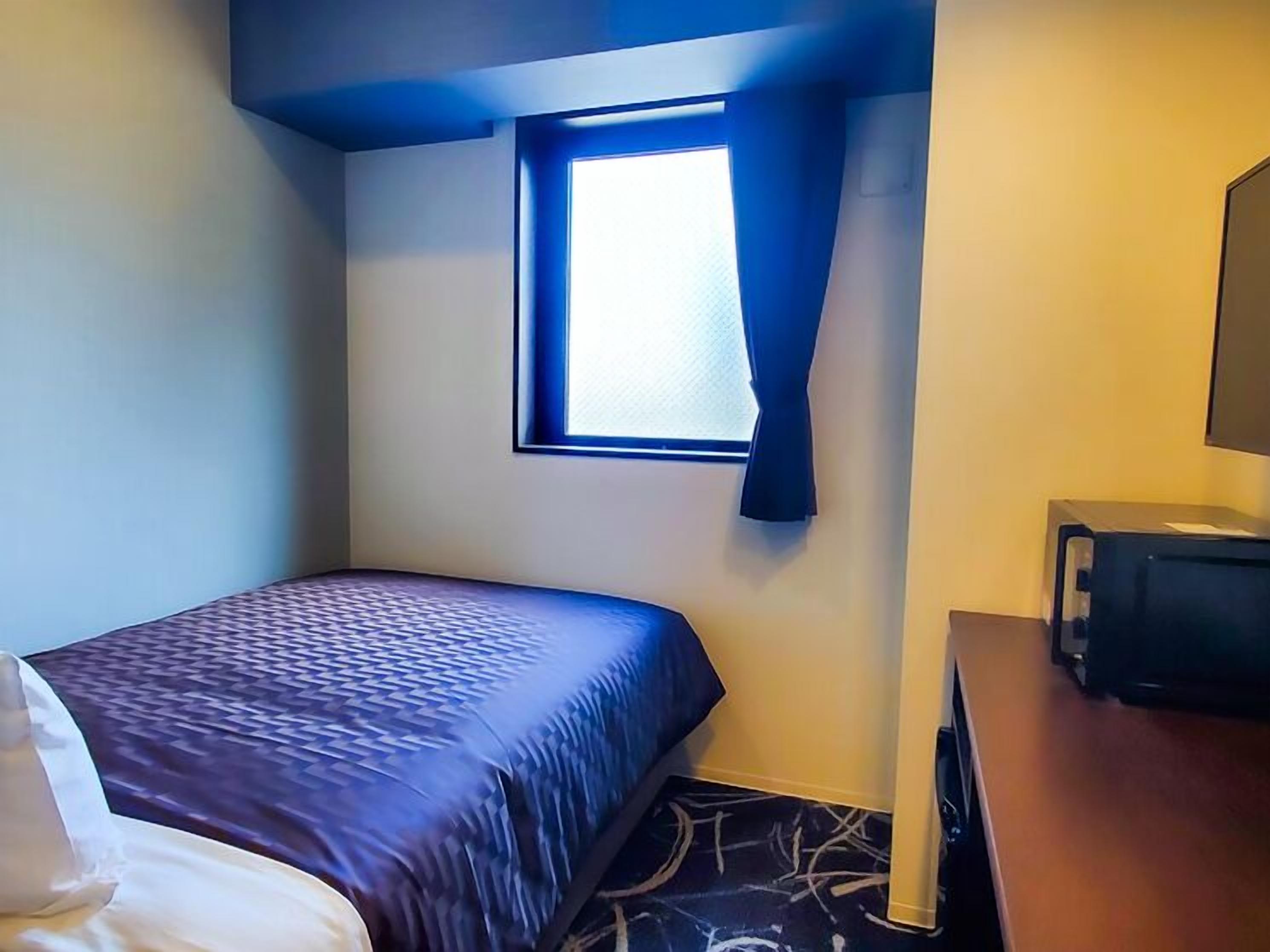Une petite chambre d’hôtel avec un lit simple, une fenêtre avec des rideaux, un bureau avec un ordinateur et un tapis à motifs.
