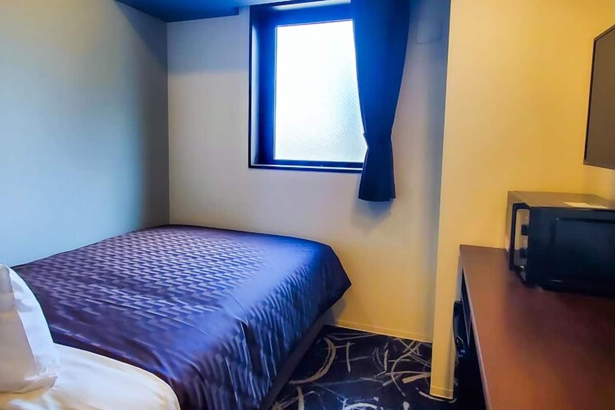 Ein kleines Hotelzimmer mit einem Einzelbett, einem fensterbelaubten Fenster, einem Schreibtisch mit Computer und einem gemusterten Teppich.
