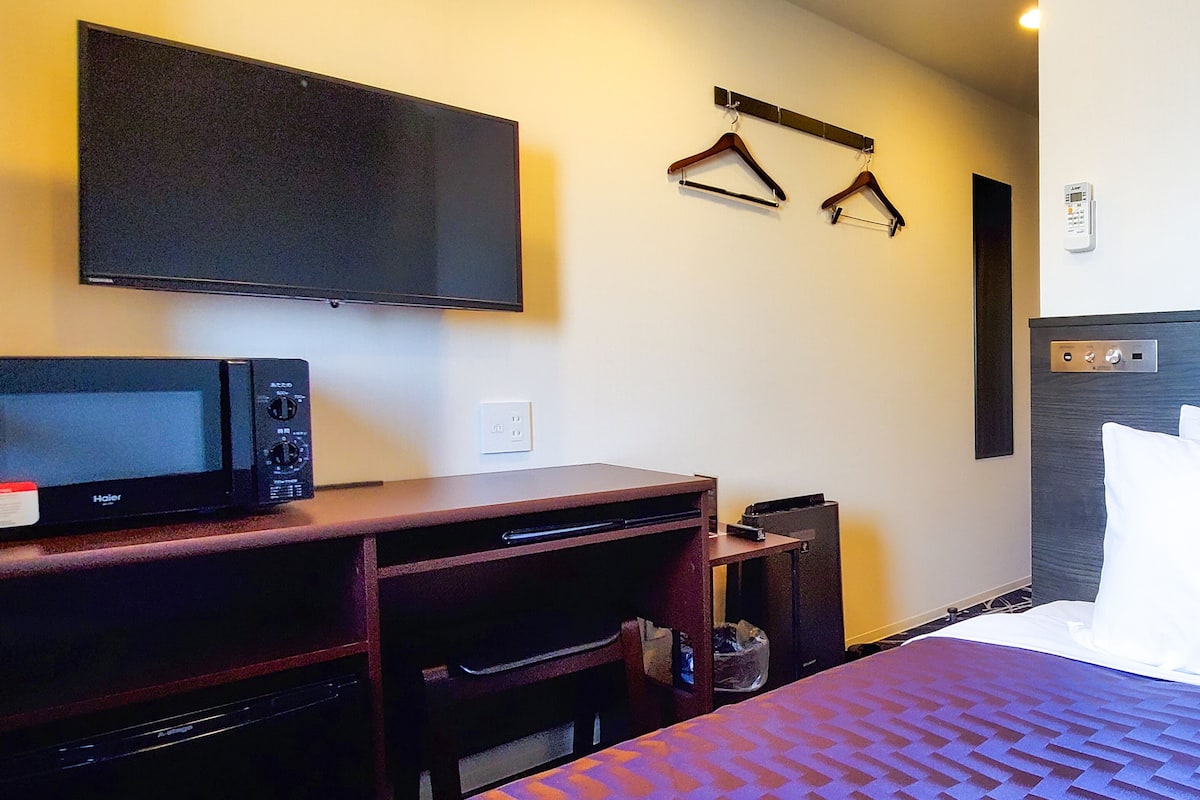 Ein Hotelzimmer mit Bett, Flachbildfernseher an der Wand, einer Mikrowelle, einem kleinen Tisch und einem Kleiderständer mit Kleiderbügeln.
