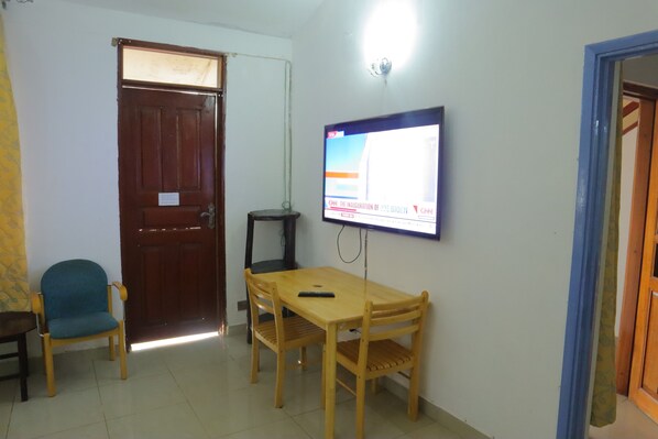 A small room with a wooden table, two chairs, a television mounted on the wall, and a door. - Kogelo Suites (Kisumu)