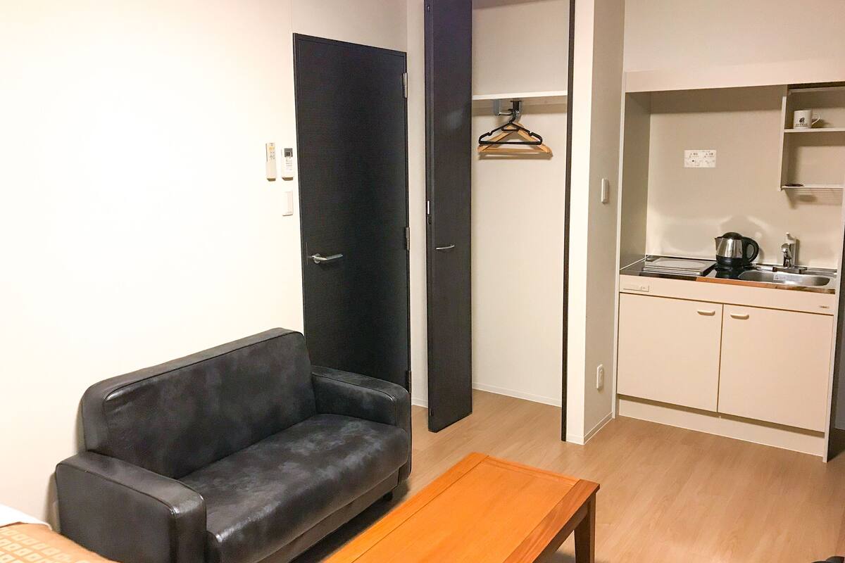 A compact living space with a sofa, a wooden table, a kitchenette, and a wardrobe.