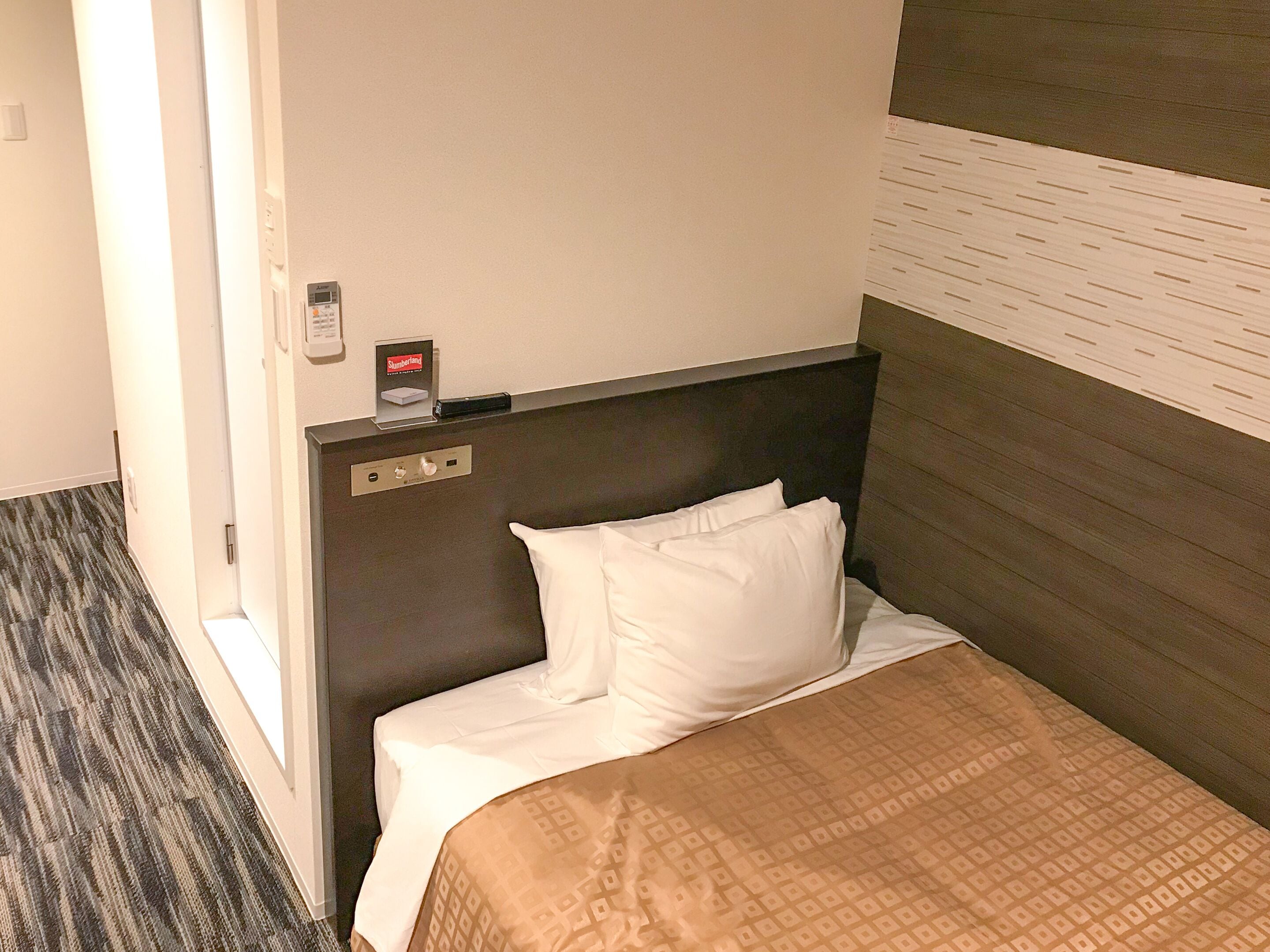A hotel room with a bed, a headboard, a control panel, and a wall-mounted air conditioning unit.