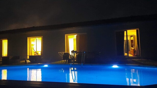 A nighttime swimming pool with three open doors leading to interior spaces, each illuminated by warm interior lighting.