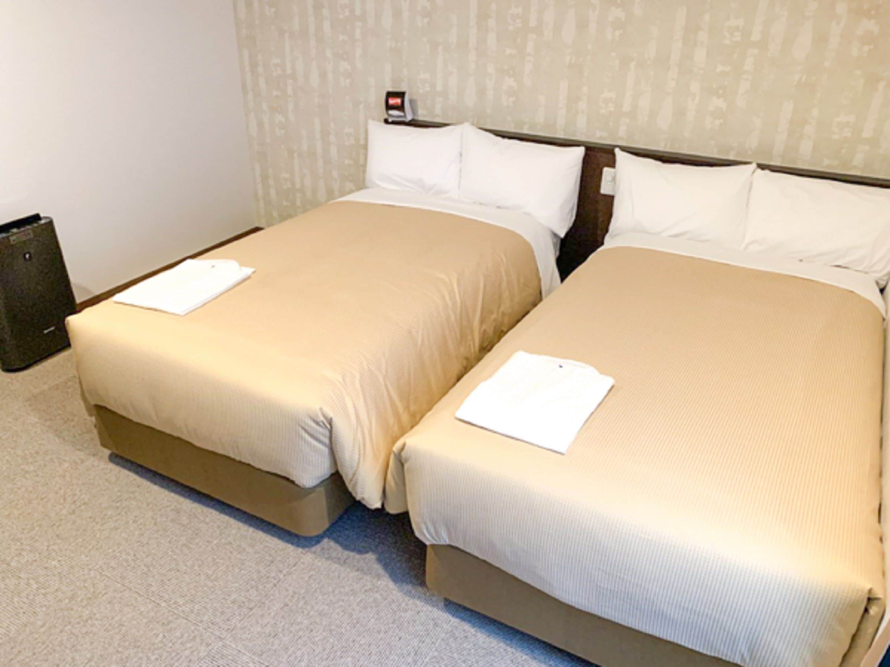 A hotel room with two beds, each with a beige bedspread and white pillows. There is a small table with a lamp and a wall-mounted clock.
