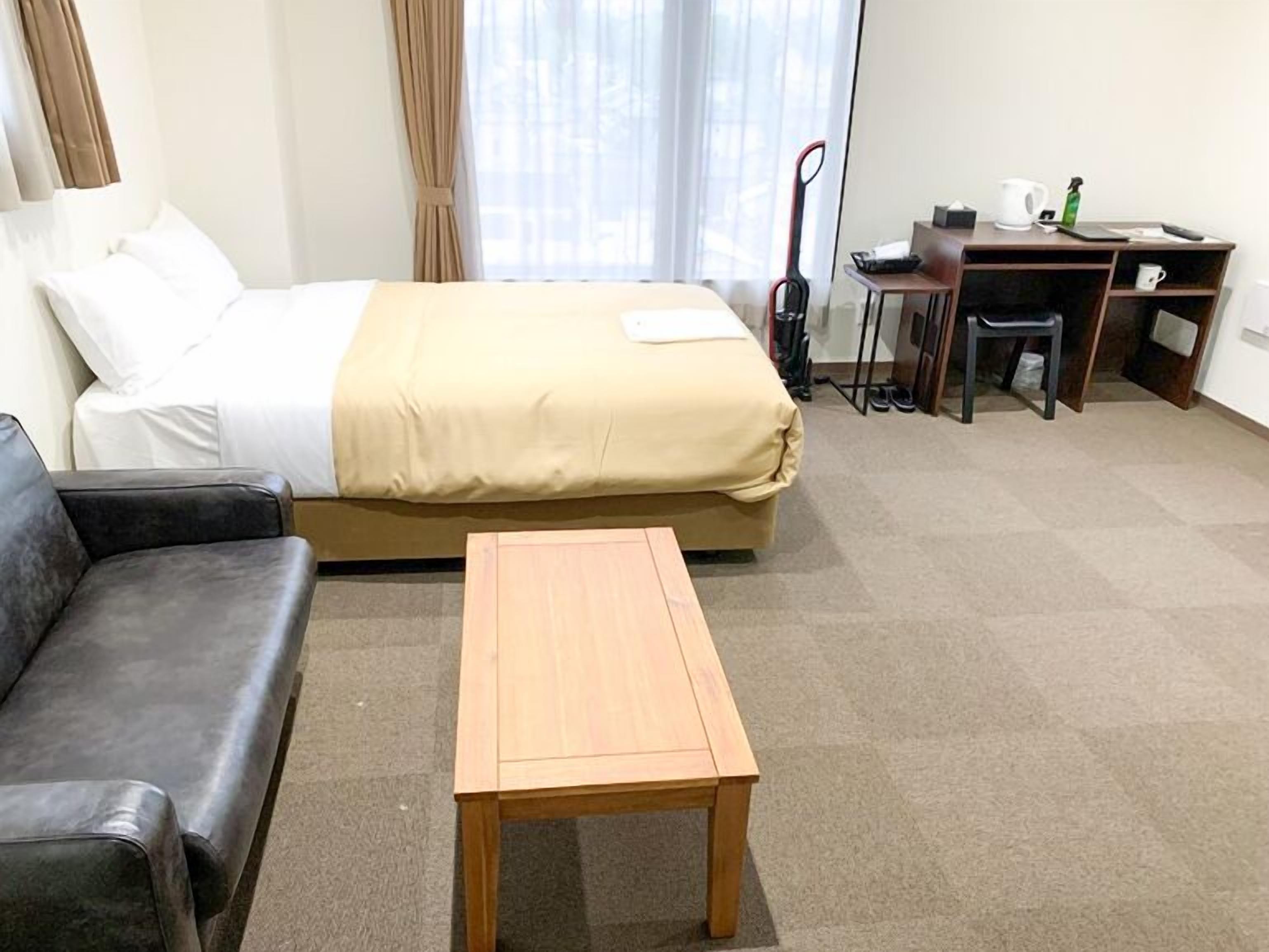 A hotel room with a bed, a wooden table, a black leather sofa, a desk with a chair, and a window with curtains.