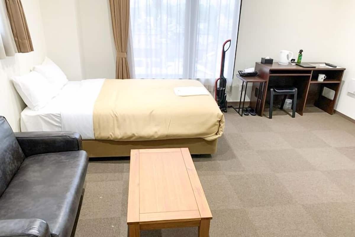 A hotel room with a bed, a wooden table, a black leather sofa, a desk with a chair, and a window with curtains.