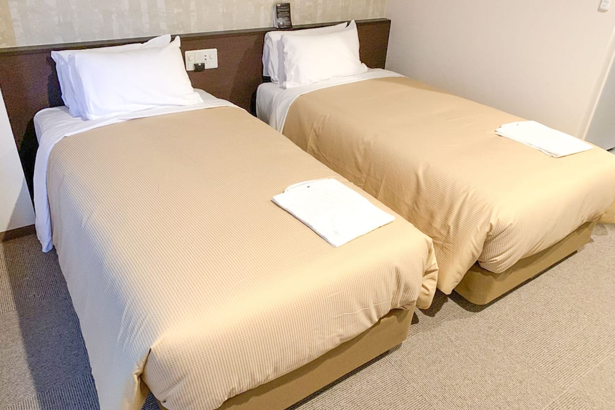 A hotel room with two single beds, each with a beige bedspread and white pillows.