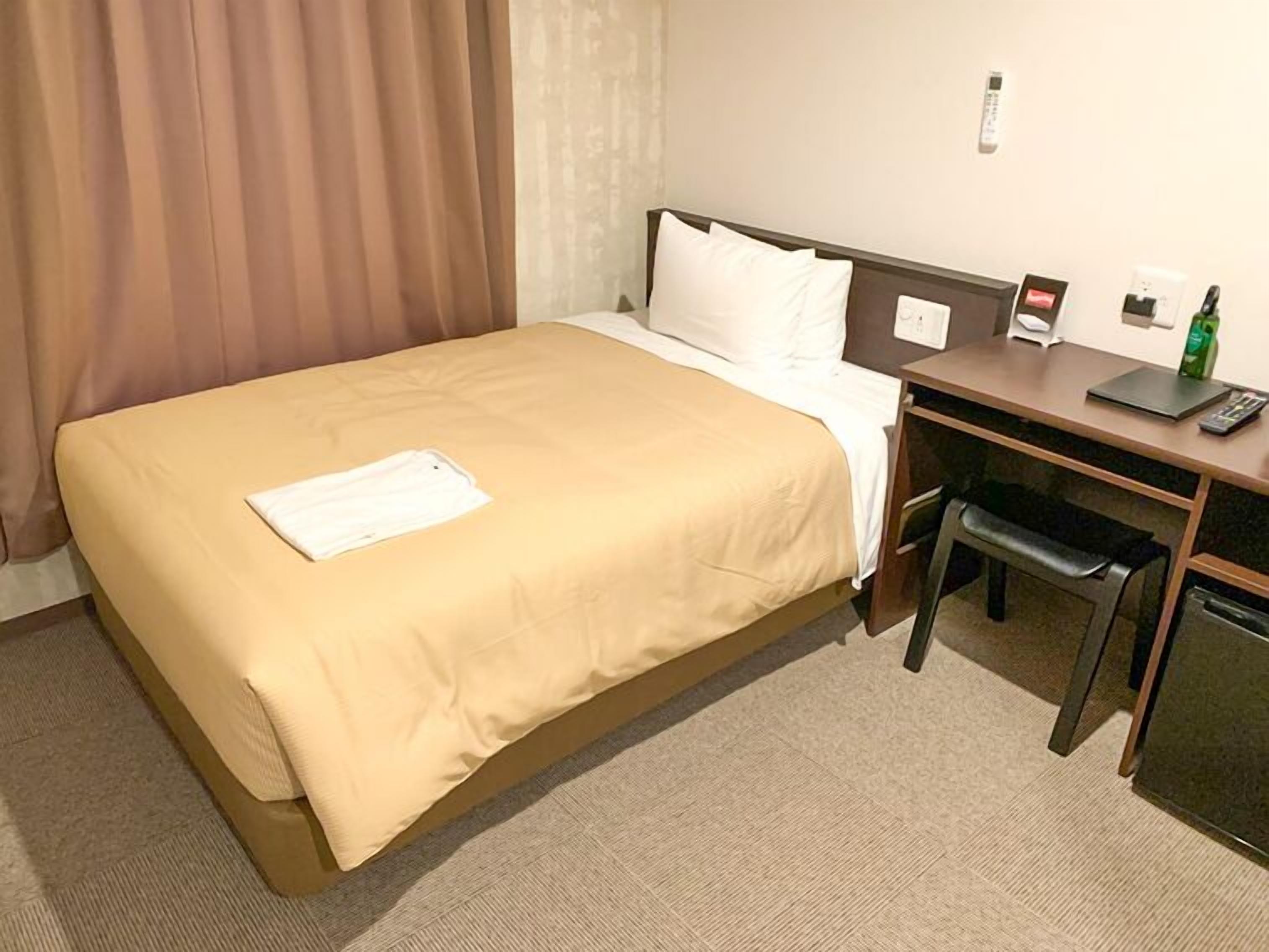 A hotel room with a bed, a desk, a chair, a desk lamp, a telephone, and a small table with a bottle.