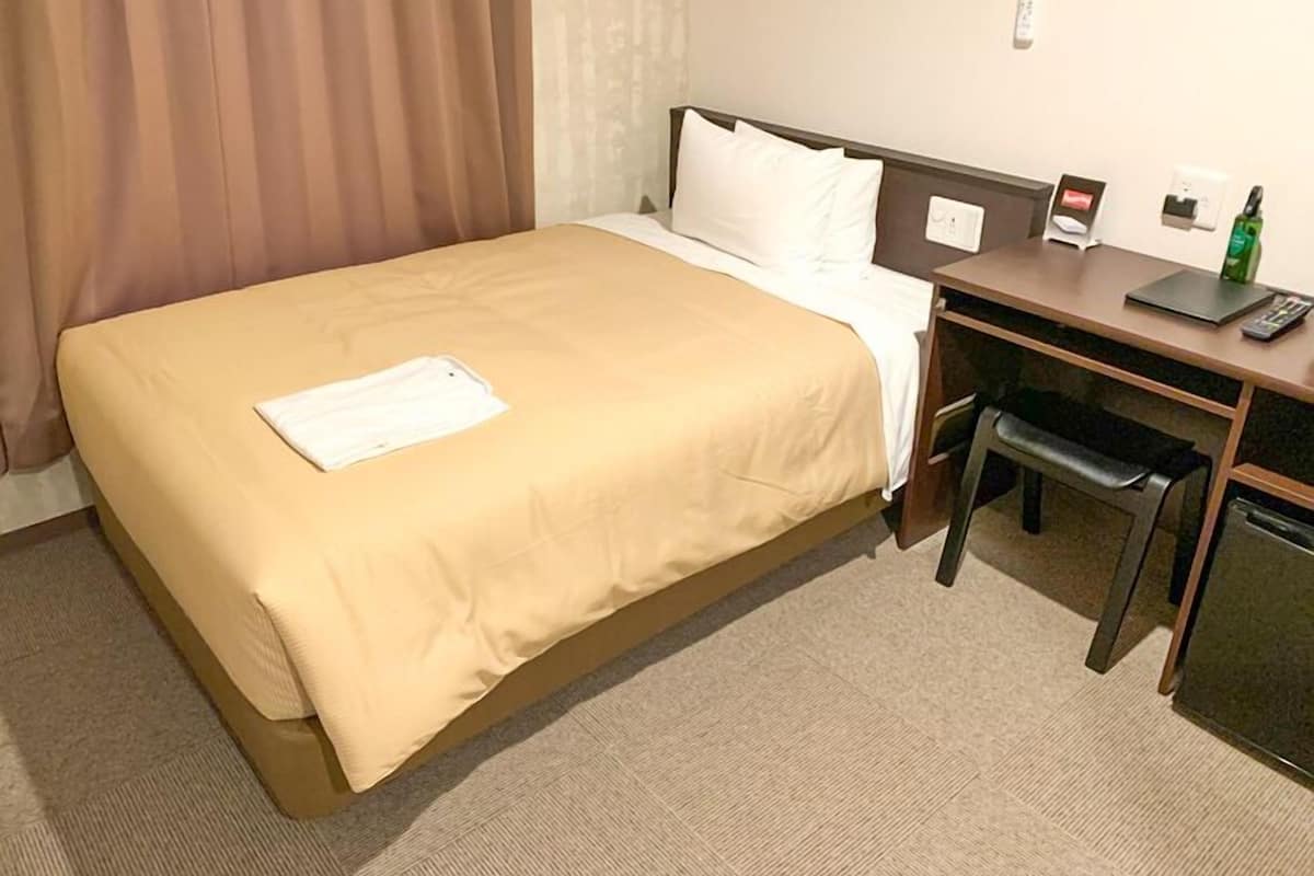 A hotel room with a bed, a desk, a chair, a desk lamp, a telephone, and a small table with a bottle.