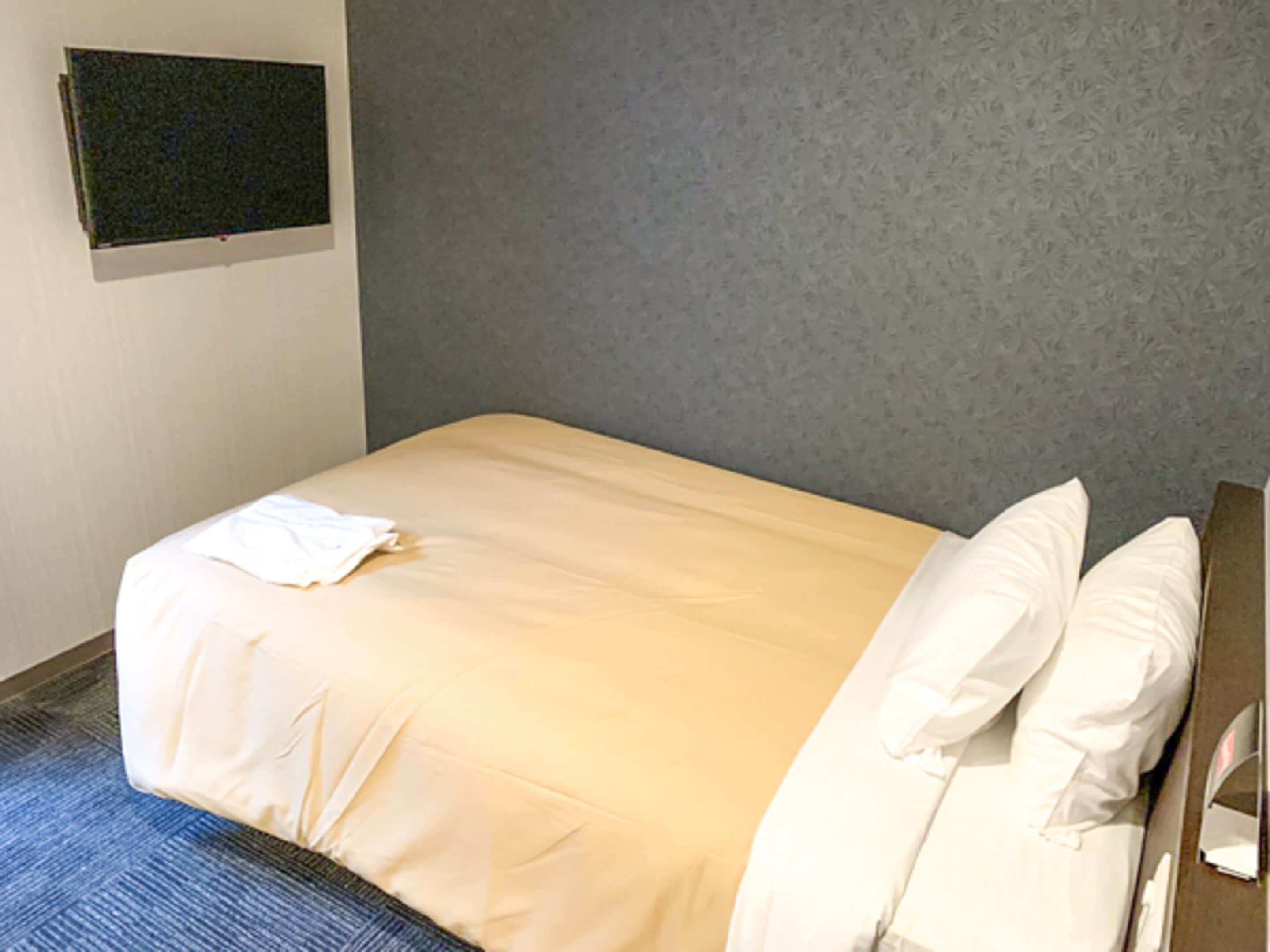 A single bed with white linens in a room with a grey textured wall and a blue carpet.