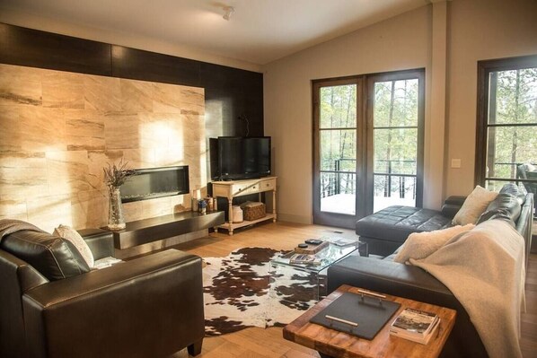 A modern living room with a fireplace, leather sofas, a glass coffee table, and large windows offering a view of trees.