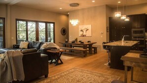 A spacious living area with a dark leather sofa, a wooden coffee table, and a dining table with benches. There is a chandelier above the dining area and a kitchen with dark cabinets and stainless steel appliances. - Gleneagles Golf Course Retreat with Hot Tub (Columbia Falls)