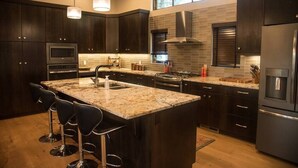 A modern kitchen with dark wood cabinets, stainless steel appliances, a large island with bar stools, and pendant lighting. - Gleneagles Golf Course Retreat with Hot Tub (Columbia Falls)