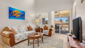 Ein Wohnzimmer mit einer Couch, zwei Sesseln, einem Couchtisch und einem Fernseher. An der Wand hängt ein Gemälde einer Meeresschildkröte, es gibt eine Lampe und einen Blick auf einen Balkon mit Geländer.