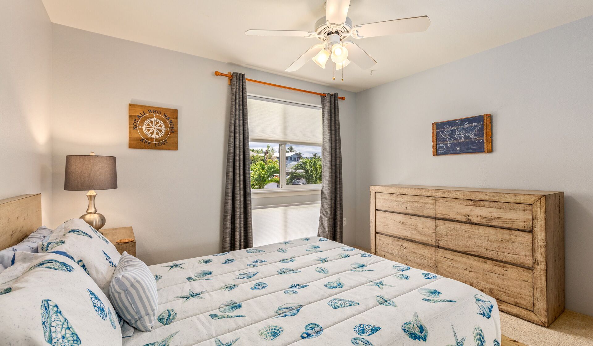 A bedroom with a bed, a wooden dresser, a ceiling fan, a window with curtains, and two wall decorations.