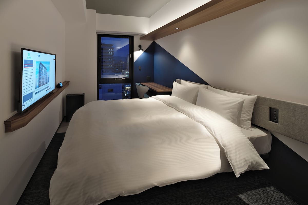 A modern hotel room with a large bed, a TV mounted on a wooden shelf, a view of the cityscape through a glass door, and a dark-colored carpet.