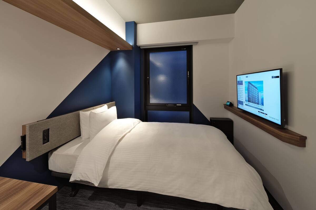 A modern hotel room with a bed, a desk, a TV mounted on the wall, and a window with a view of a building.