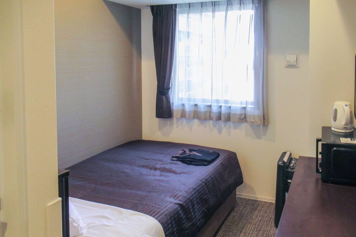 Une petite chambre d’hôtel avec un lit simple, un bureau avec une bouilloire et une fenêtre avec des rideaux.