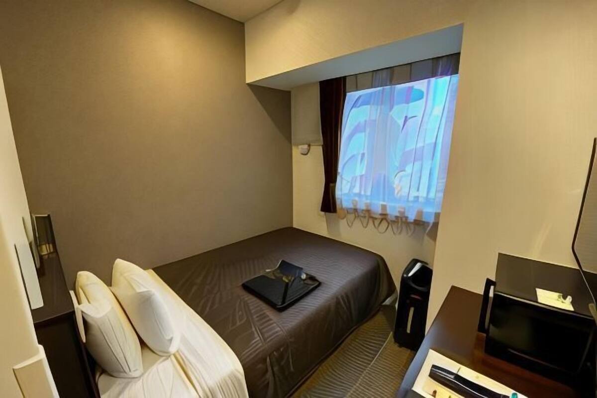 Une chambre d’hôtel avec un lit simple, un bureau avec un ordinateur, une chaise et une fenêtre avec des rideaux.