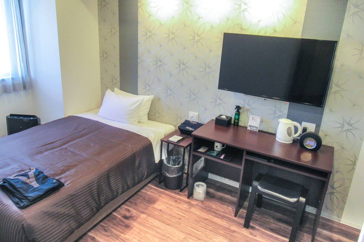 Une chambre d’hôtel équipée d’un lit, d’un bureau, d’une chaise, d’une télévision, ainsi que d’une petite table sur laquelle se trouvent une bouilloire et une poubelle.