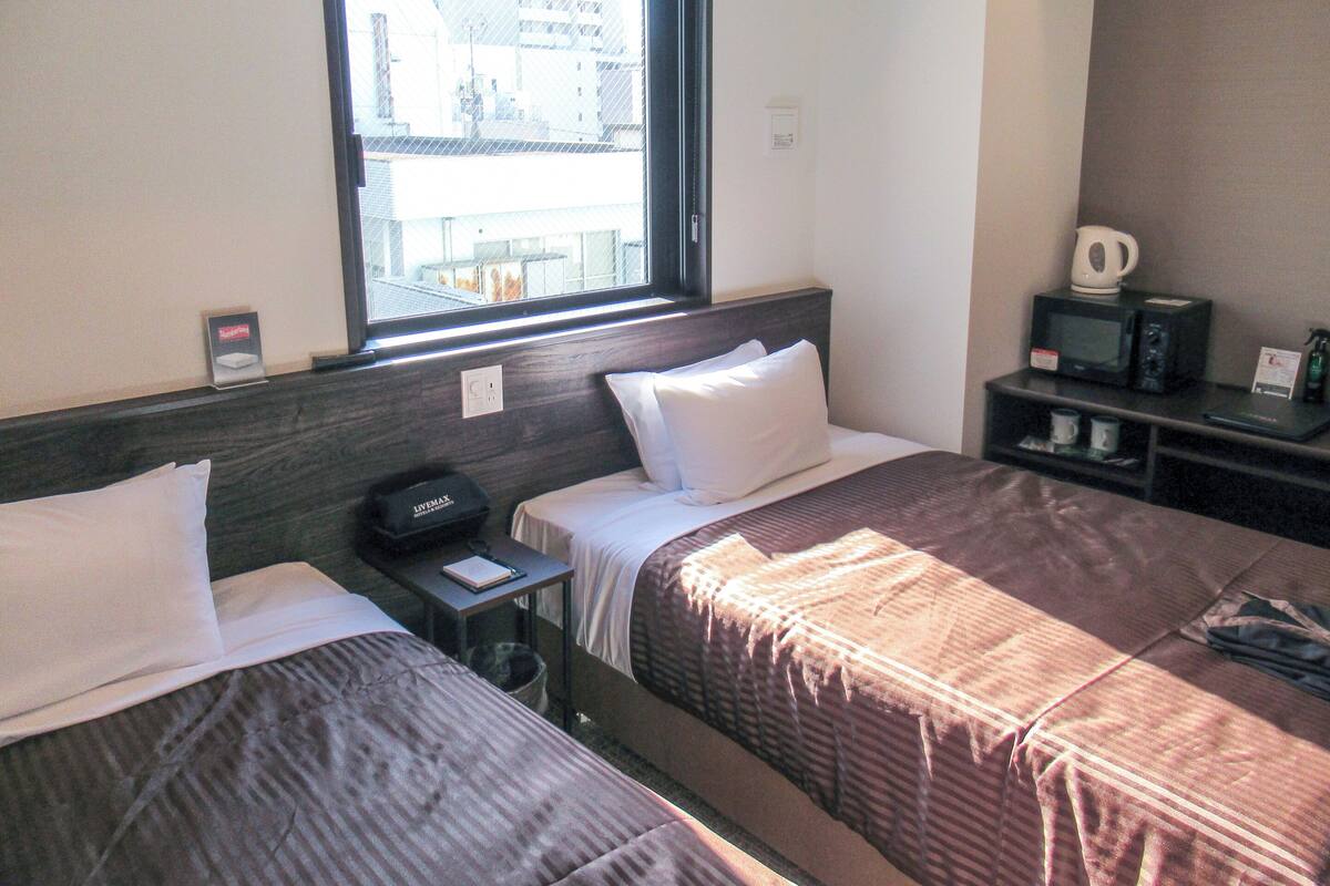Une chambre d’hôtel avec deux lits, une fenêtre donnant sur des immeubles, un bureau avec un micro-ondes et une petite poubelle.