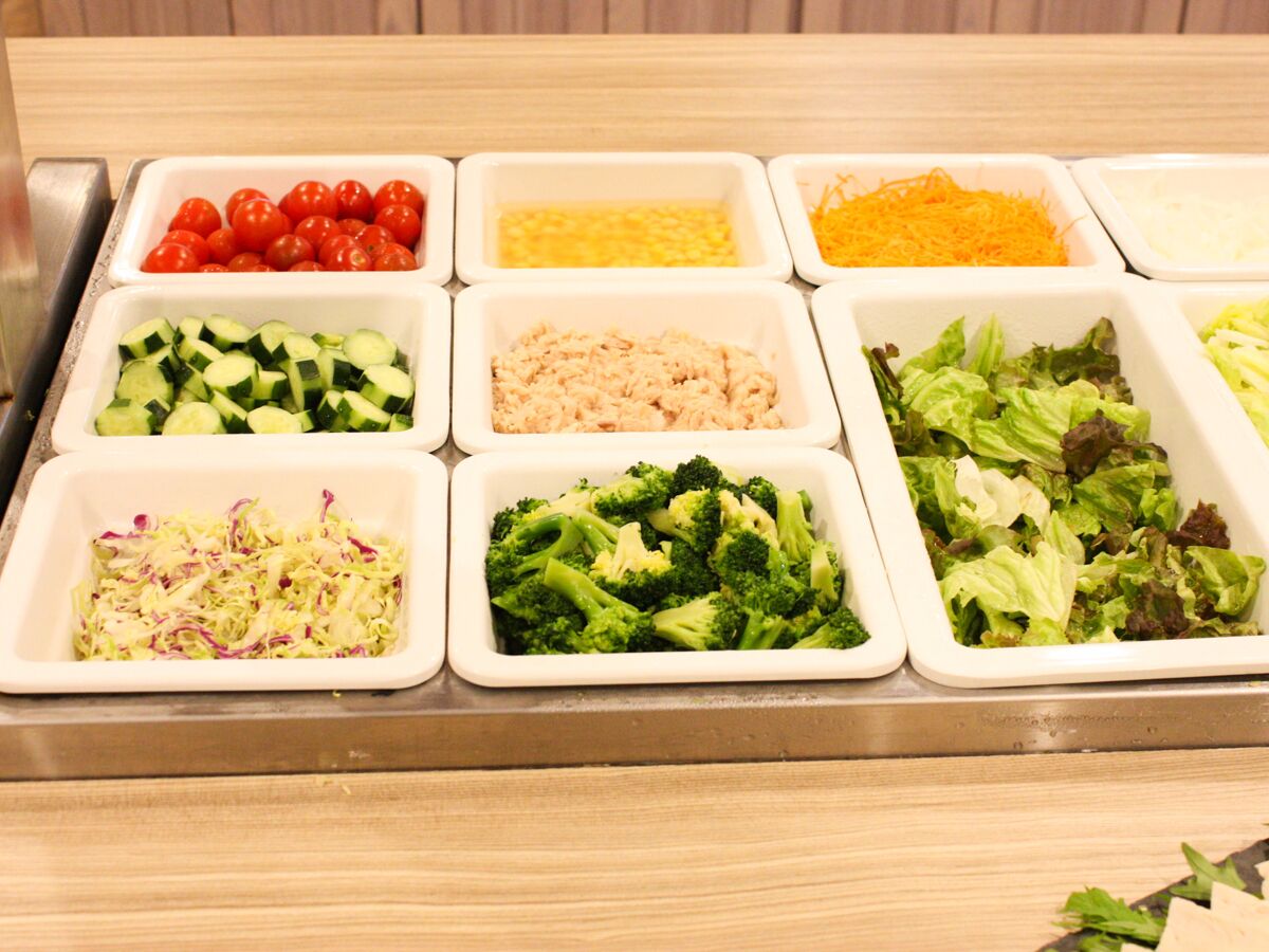 A buffet with various food items including tomatoes, cucumbers, shredded cheese, shredded carrots, broccoli, and mixed greens.