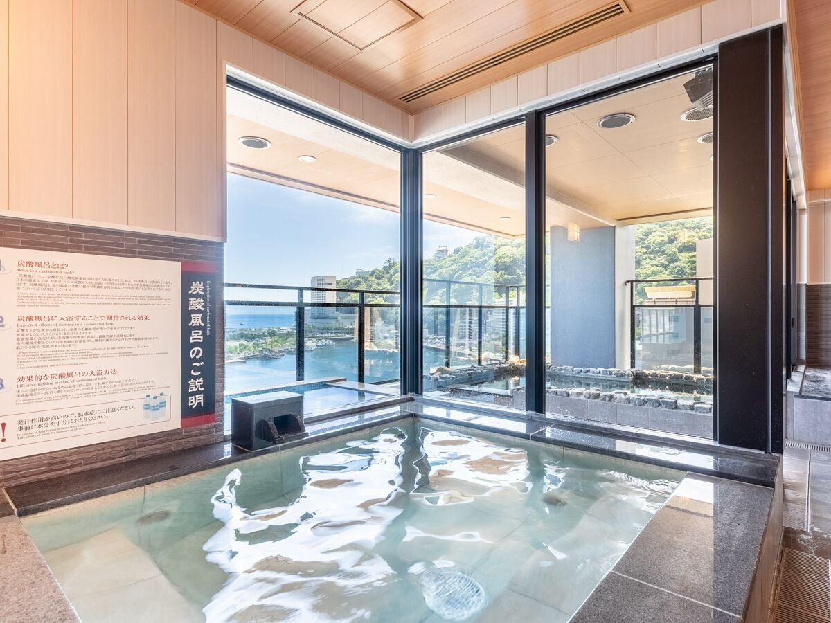 A modern indoor swimming pool with clear glass walls, a view of the sea, and a wooden ceiling.