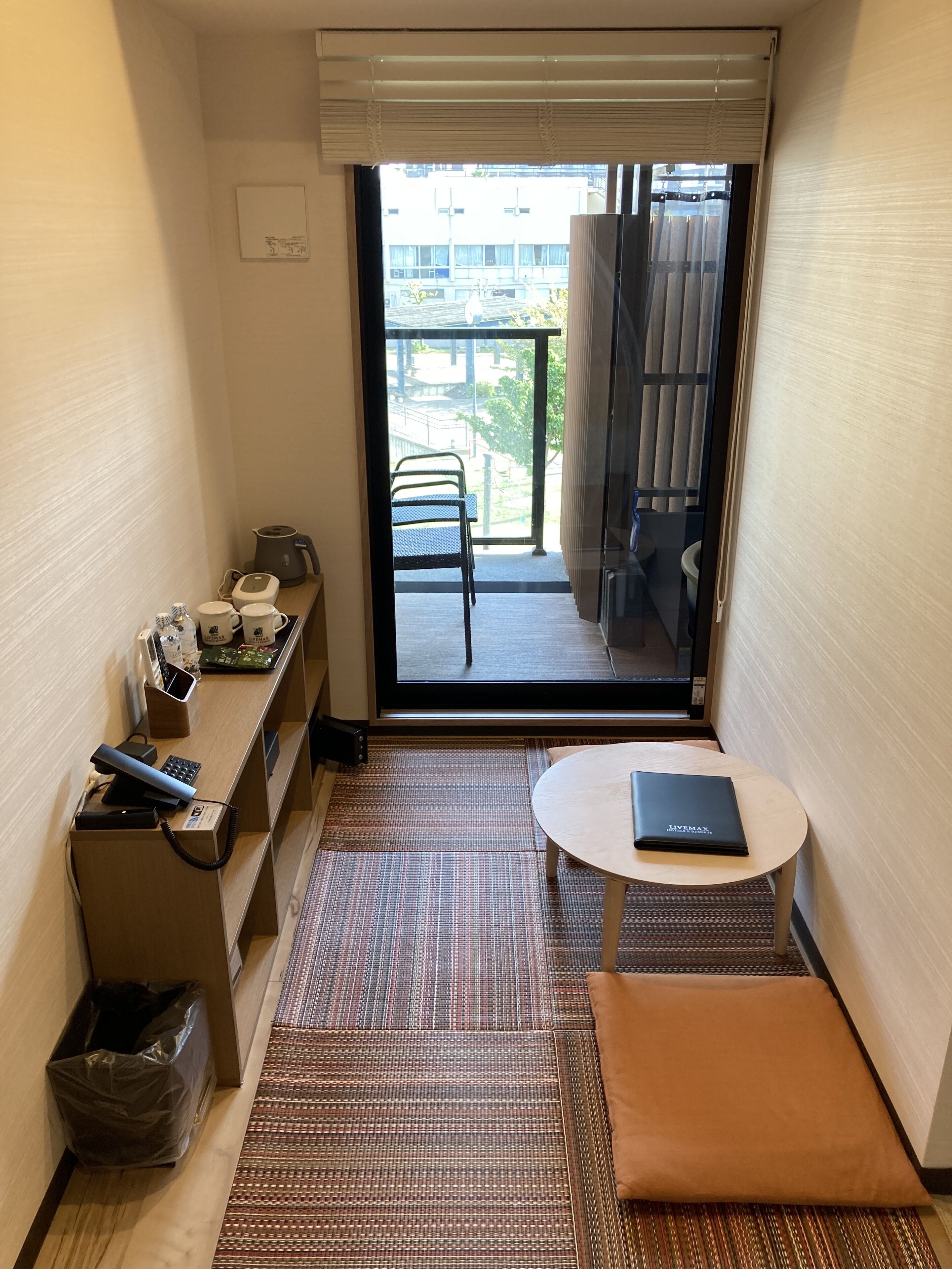 A compact hotel room with a small desk, a chair, a table, a sofa, and a view of the outside balcony.