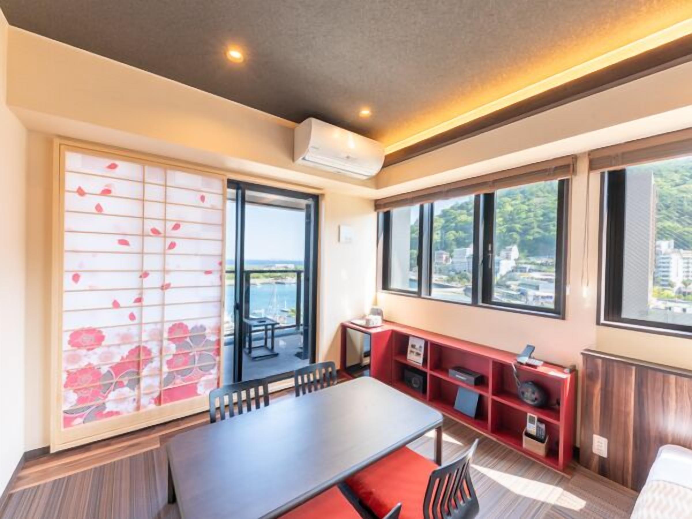 A hotel room with a dining table, chairs, a red bookshelf, a sliding door with a floral pattern, and large windows offering a view of the sea and buildings.