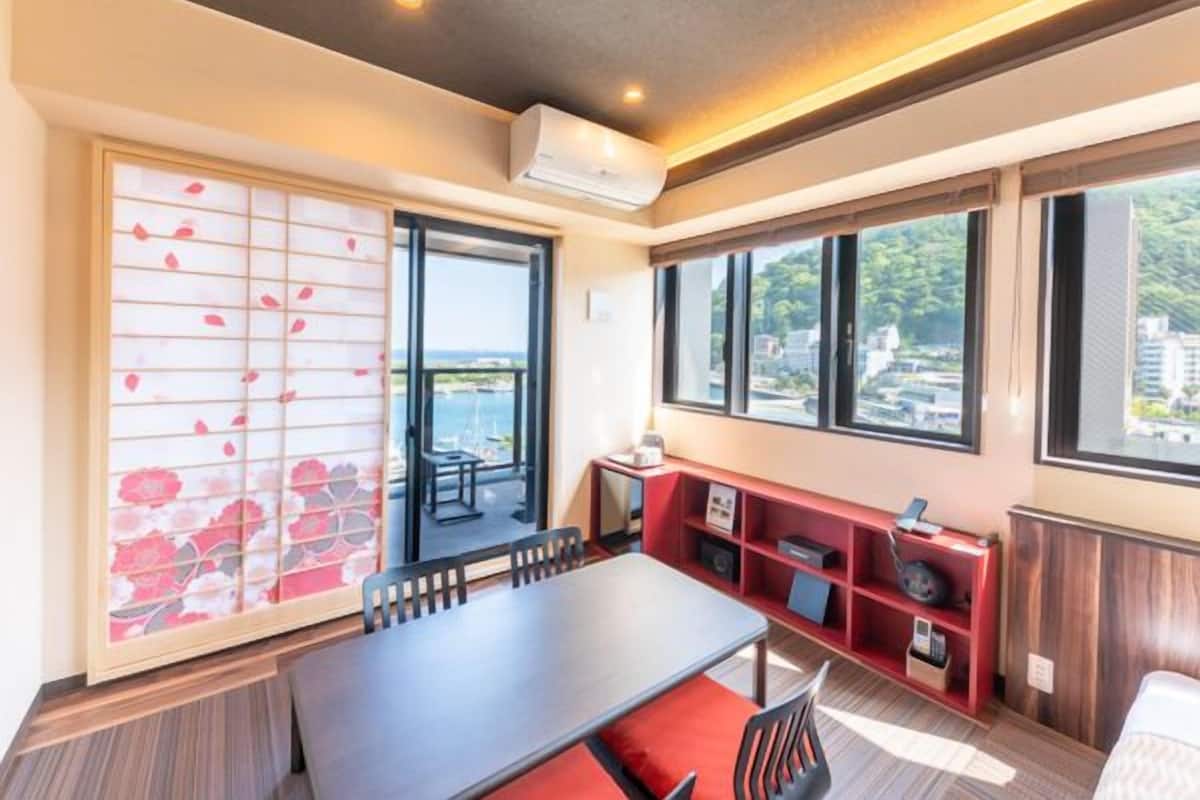 A hotel room with a dining table, chairs, a red bookshelf, a sliding door with a floral pattern, and large windows offering a view of the sea and buildings.