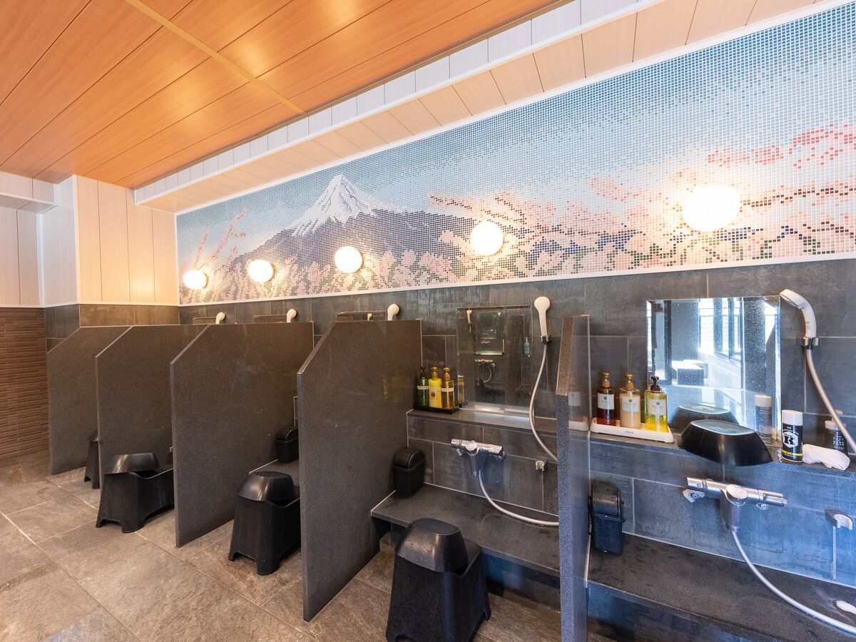 Baño público con cabinas individuales, un espejo grande y un lavamanos de pared con jabón de manos y champú.