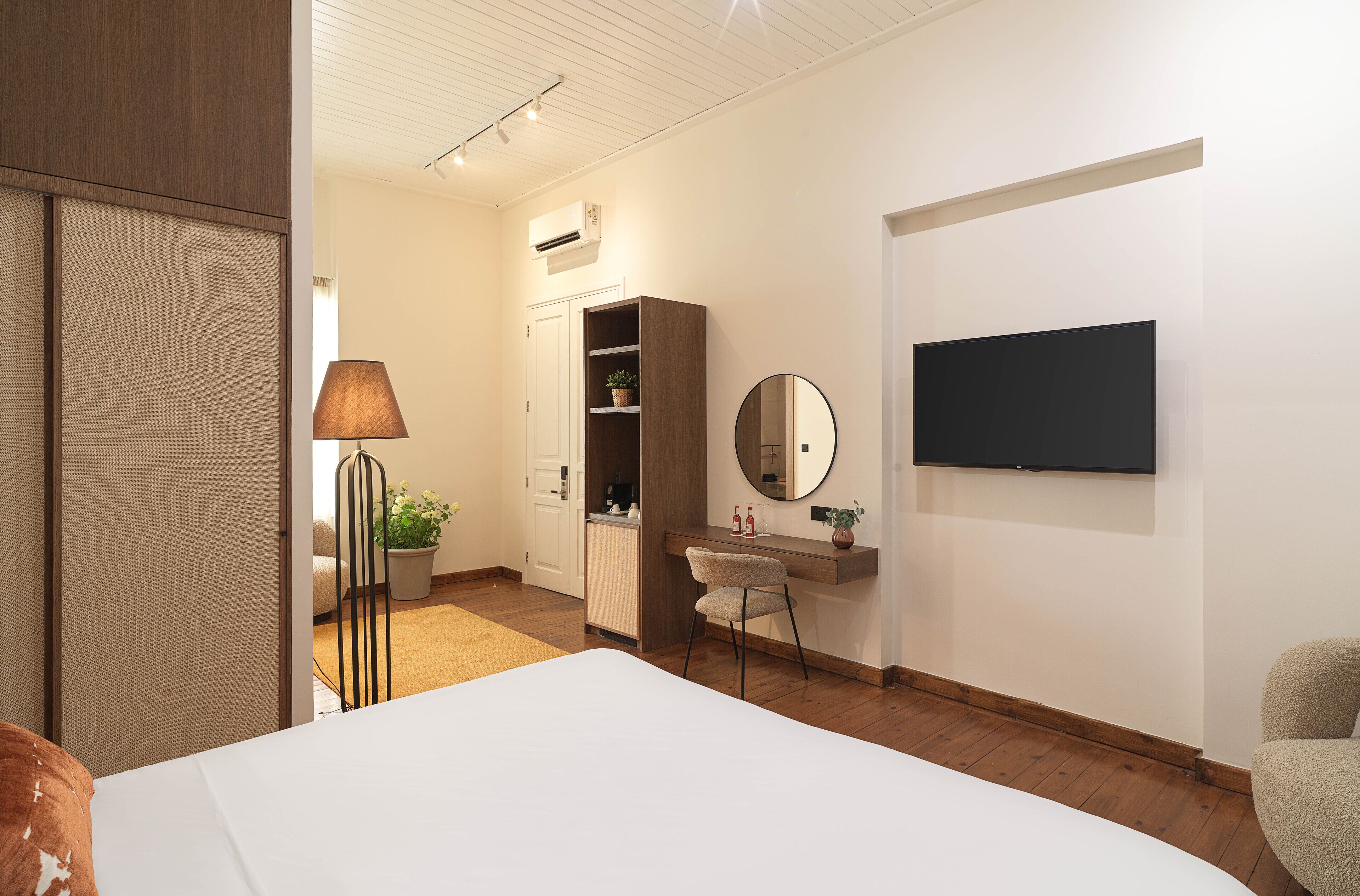 A modern hotel room with a bed, a desk, a chair, a lamp, a mirror, and a TV on the wall.